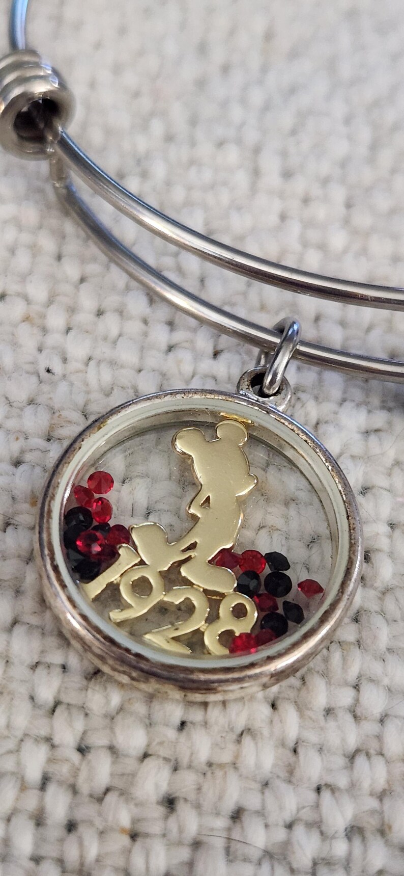 Puede incluir: Pulsera de brazalete plateada con un dije redondo. El dije presenta una silueta dorada de Mickey Mouse y el a&ntilde;o "1928". Cristales rojos y negros est&aacute;n dentro del dije. La pulsera est&aacute; sobre una superficie blanca texturizada.