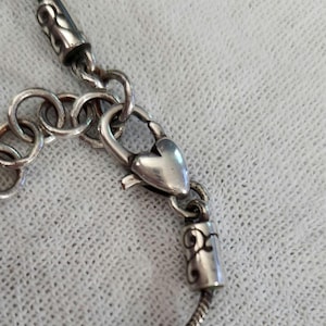 May include: Close-up of a silver necklace with a heart-shaped clasp. The necklace has a snake chain design and decorative cylindrical end pieces. The clasp and end pieces have ornate scrollwork.