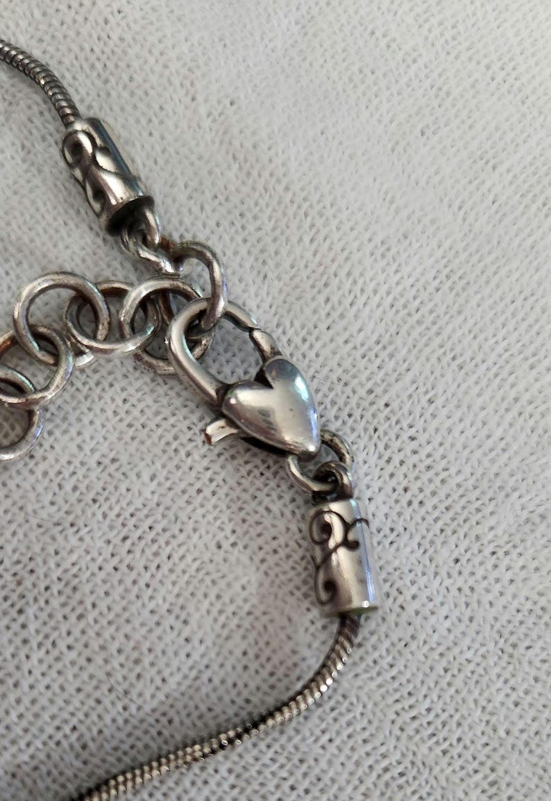 May include: Close-up of a silver necklace with a heart-shaped clasp. The necklace has a snake chain design and decorative cylindrical end pieces. The clasp and end pieces have ornate scrollwork.