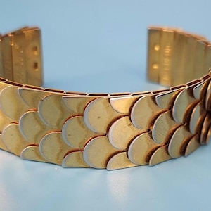May include: A gold-toned bracelet with a scale-like design. The bracelet is made of interlocking, fan-shaped metal pieces. The bracelet is open at the ends and has a light blue background.