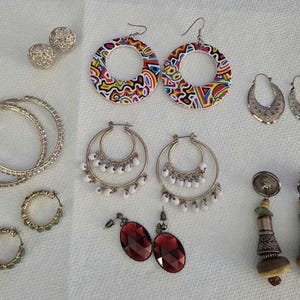 May include: A collection of various earrings including silver-tone hoops, large circular hoops with a colorful abstract pattern, crescent-shaped textured earrings, hoop earrings with white beads, oval red faceted glass drops, and tiered metal dangles.