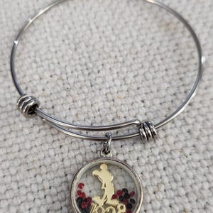 Puede incluir: Pulsera de brazalete plateada con un dije que presenta una silueta dorada de un personaje de dibujos animados y el n&uacute;mero "1928" dentro de un colgante redondo transparente. El colgante est&aacute; lleno de cuentas rojas y negras.