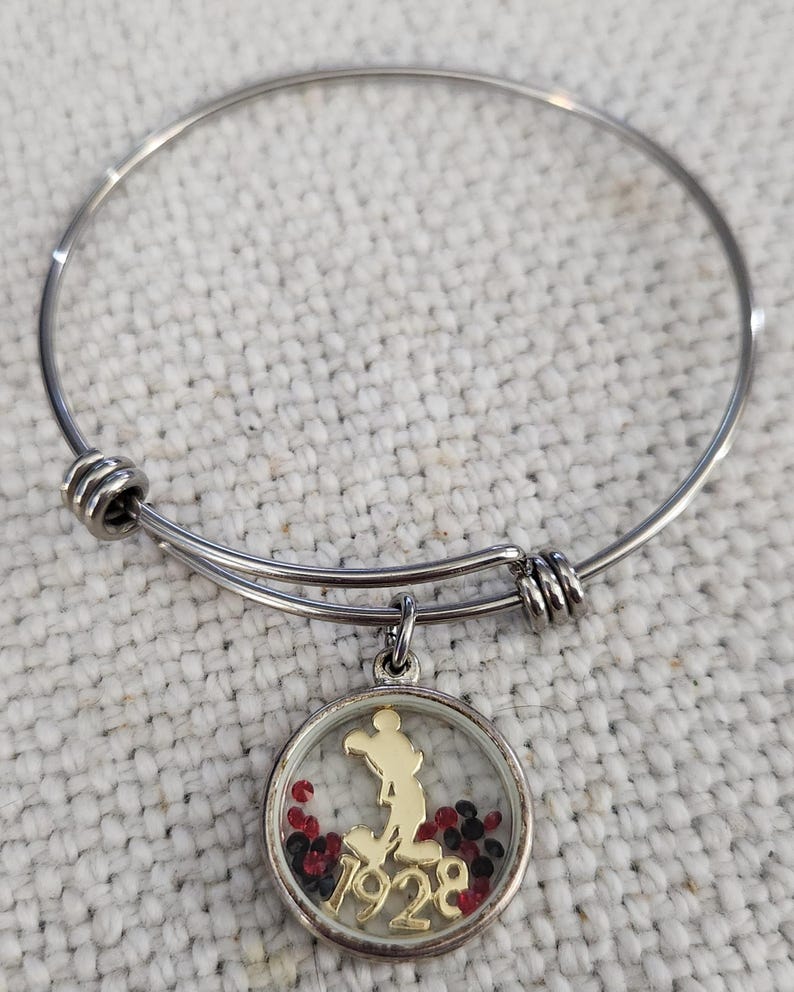 Puede incluir: Pulsera de brazalete plateada con un dije que presenta una silueta dorada de un personaje de dibujos animados y el n&uacute;mero "1928" dentro de un colgante redondo transparente. El colgante est&aacute; lleno de cuentas rojas y negras.