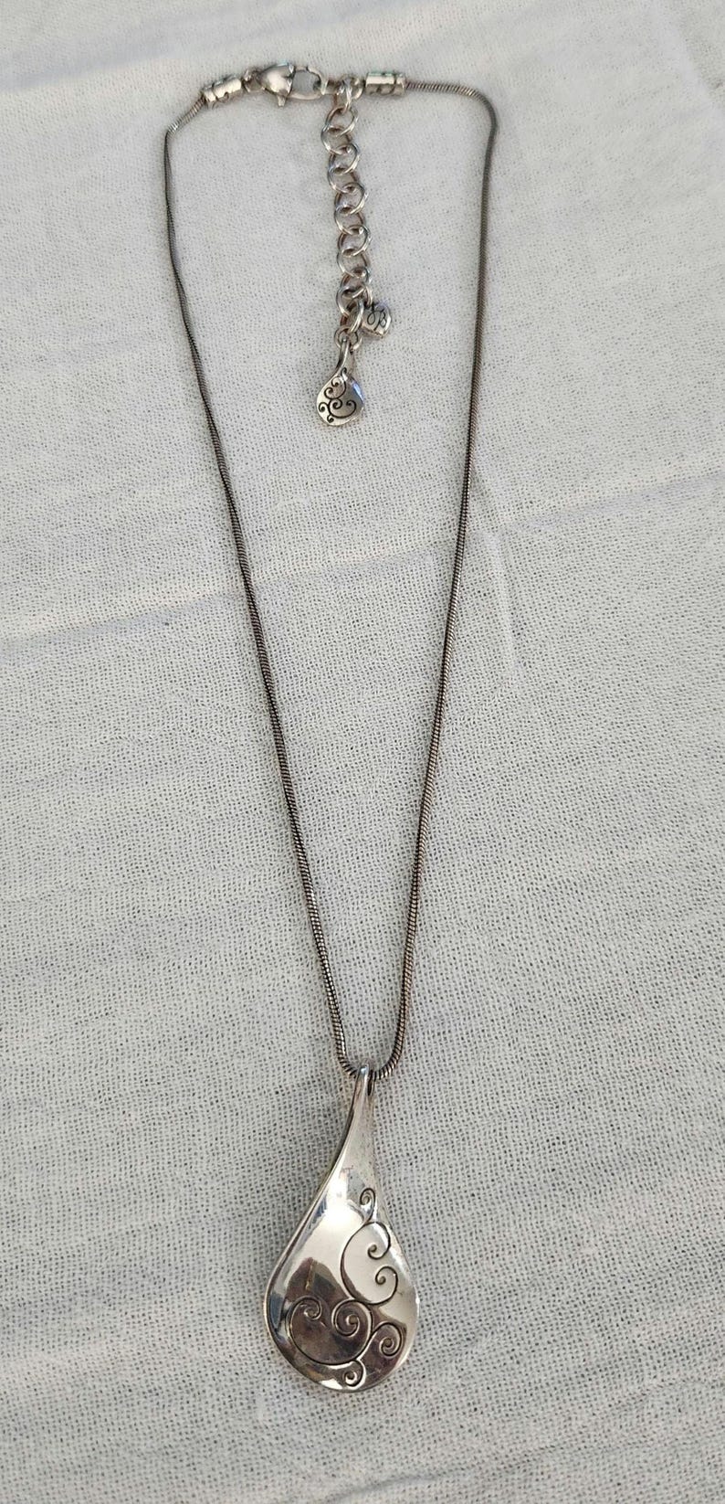 May include: Silver-toned necklace with a teardrop pendant. The pendant features an engraved swirl design. The necklace has a delicate chain and an adjustable length. A small charm hangs near the clasp.