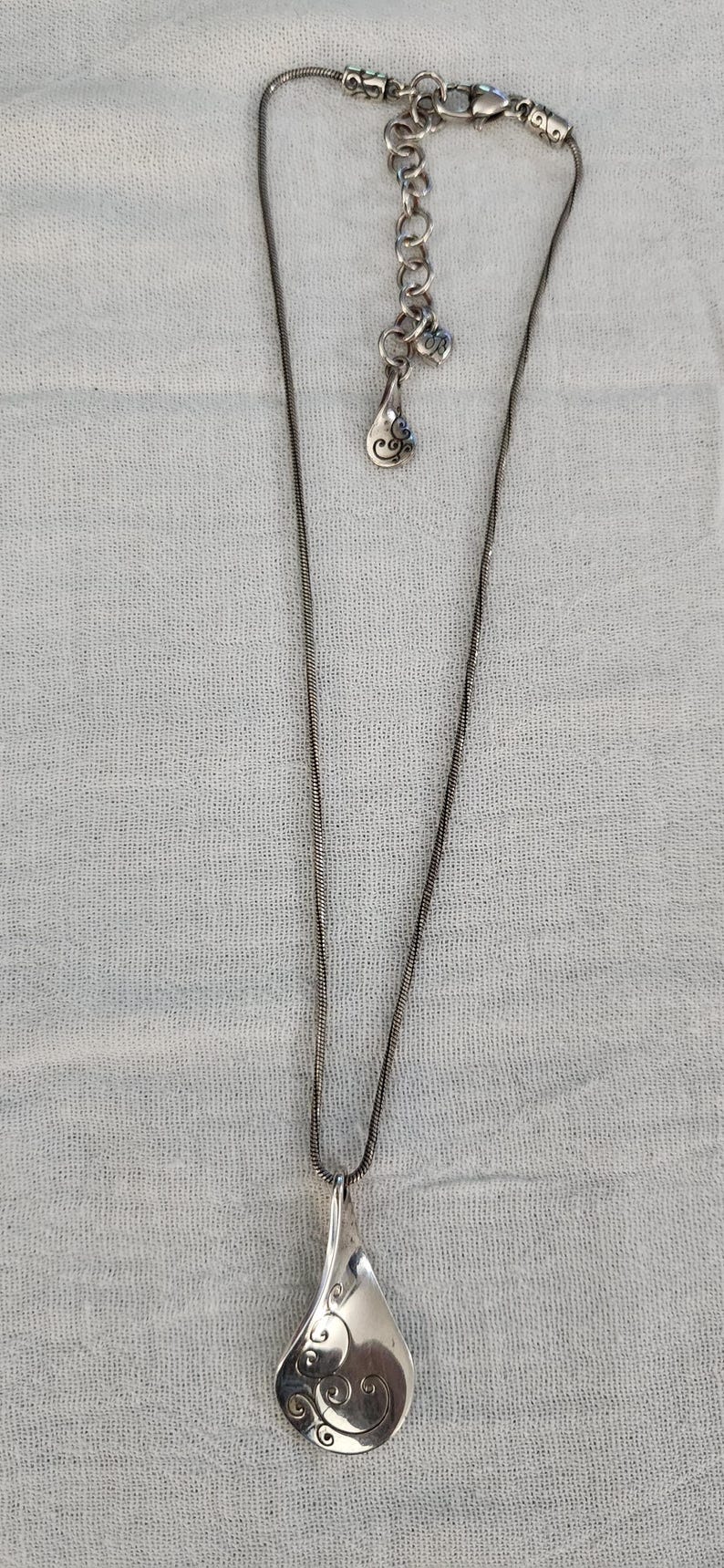 May include: Silver-toned necklace with a teardrop pendant. The pendant features an etched swirl design. The necklace has an adjustable chain and a small charm near the clasp. The background is a light gray fabric.