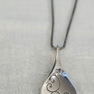 May include: A silver-toned pendant necklace with a teardrop-shaped pendant. The pendant features an etched, swirling design on its surface. The necklace chain is a dark silver color, and the background is a white, textured fabric.