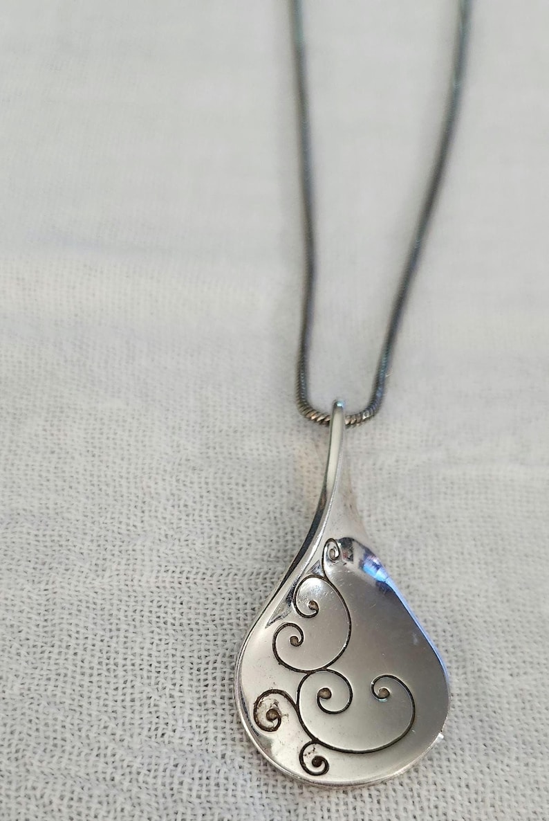 May include: A silver-toned pendant necklace with a teardrop-shaped pendant. The pendant features an etched, swirling design on its surface. The necklace chain is a dark silver color, and the background is a white, textured fabric.