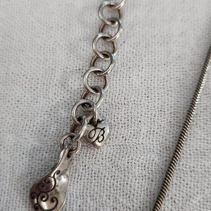 May include: Silver-toned necklace with a decorative pendant. The pendant features a heart-shaped charm with the letter "B" and a teardrop-shaped element with an ornate design. The necklace has a delicate chain.