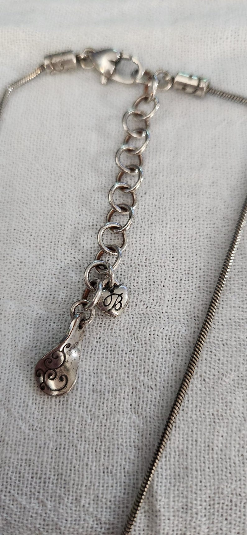 May include: Silver-toned necklace with a decorative pendant. The pendant features a heart-shaped charm with the letter "B" and a teardrop-shaped element with an ornate design. The necklace has a delicate chain.