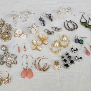 May include: A collection of various earrings, including gold hoops, pearl studs, and drop earrings with pink stones. Other styles include floral, bow, and geometric designs. The earrings are displayed on a white surface.