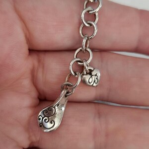 May include: A silver-toned charm bracelet with a decorative spoon-shaped charm and a heart-shaped charm. The spoon charm has an ornate, swirling design. The heart charm features the letter "B". The bracelet is made of linked circles.