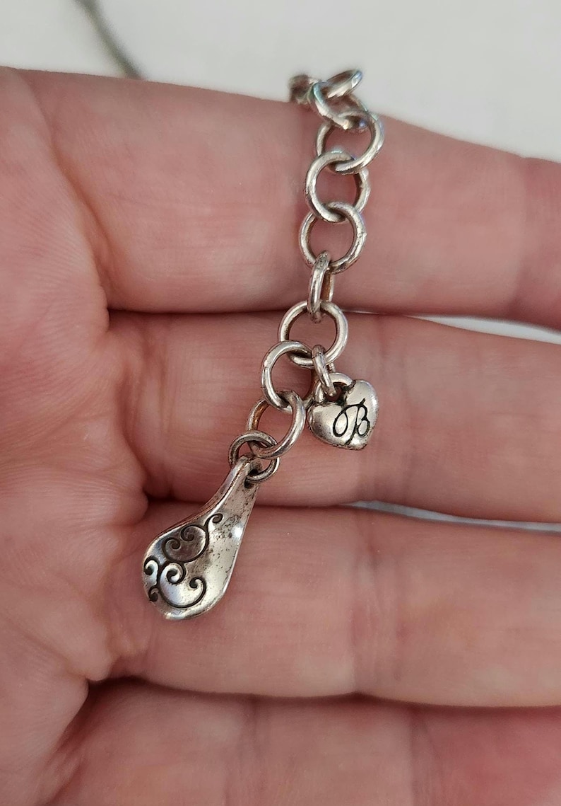 May include: A silver-toned charm bracelet with a decorative spoon-shaped charm and a heart-shaped charm. The spoon charm has an ornate, swirling design. The heart charm features the letter "B". The bracelet is made of linked circles.