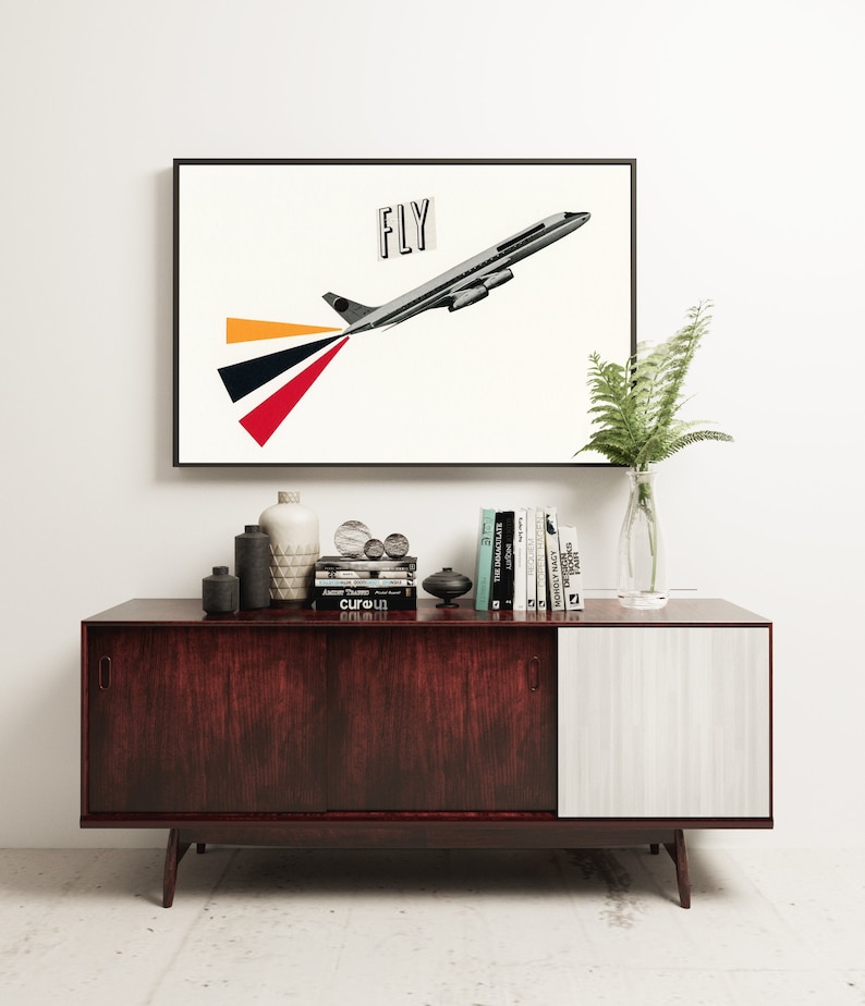 May include: A framed print of a vintage airplane with the word "FLY" in black text on a white background. The airplane is gray and has red, orange, and black stripes coming out of the back. The print is sitting on a dark wood and white cabinet with books and decorative objects.