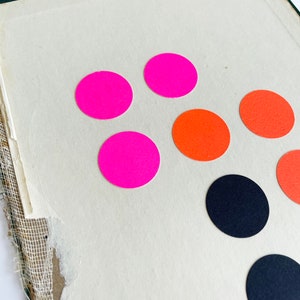 K&ouml;nnte beinhalten: Nahaufnahme einer Vintage-Buchseite mit farbenfrohen runden Aufklebern. Die Aufkleber sind in leuchtendem Pink, Orange und Schwarz gehalten. Die Buchseite ist cremefarben mit zerrissenem Rand.