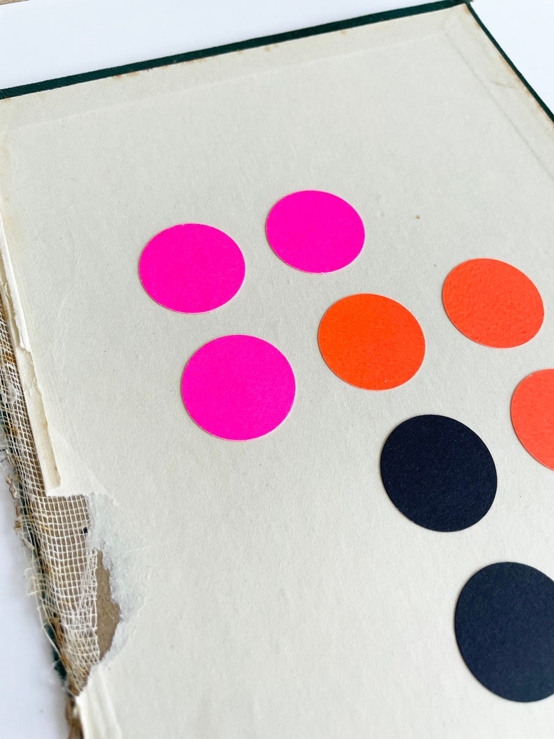 K&ouml;nnte beinhalten: Nahaufnahme einer Vintage-Buchseite mit farbenfrohen runden Aufklebern. Die Aufkleber sind in leuchtendem Pink, Orange und Schwarz gehalten. Die Buchseite ist cremefarben mit zerrissenem Rand.