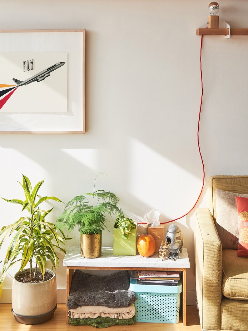 May include: A white side table with a green plant, a pumpkin, and a small rocket-shaped object. The table is next to a yellow couch with a red cord running from the wall to the table.