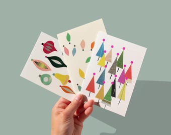 May include: A hand holds three Christmas-themed greeting cards. The cards feature colorful illustrations of ornaments, string lights, and Christmas trees. The cards have a white background and are set against a light teal backdrop.