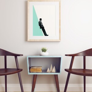 May include: A framed print of a man in a suit sitting on a light blue triangle against a white background. The print is hanging on a white wall above a light blue side table with a book and wooden geometric shapes on the shelf. Two dark brown chairs are on either side of the table.
