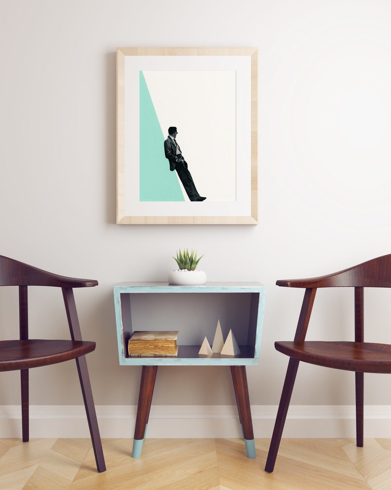 May include: A framed print of a man in a suit sitting on a light blue triangle against a white background. The print is hanging on a white wall above a light blue side table with a book and wooden geometric shapes on the shelf. Two dark brown chairs are on either side of the table.