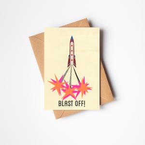 May include: A yellow card with a red, white, and blue rocket ship launching upwards. The rocket ship is surrounded by four pink and orange stars. The card says "Blast Off!" in black text.