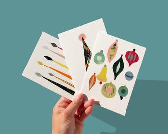 May include: Three white greeting cards with colorful illustrations are held against a teal background. The cards feature designs of paintbrushes, a party hat, and Christmas ornaments in various shapes and colors.