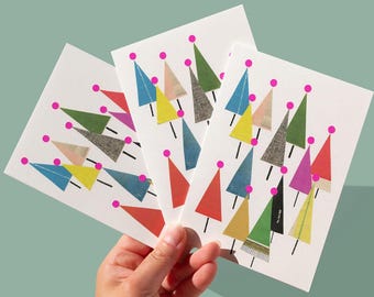 May include: A hand holds three white greeting cards with colourful geometric tree designs. Each card features a variety of triangular trees in shades of red, green, yellow, and blue, topped with pink circles. The cards are set against a light teal background.