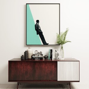 May include: A mid-century modern sideboard with a dark wood finish and a white door. The sideboard is decorated with a framed print of a man in a suit standing against a teal background, a vase of ferns, and a collection of books and decorative objects.