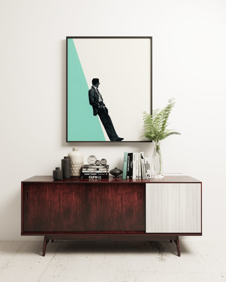 May include: A mid-century modern sideboard with a dark wood finish and a white door. The sideboard is decorated with a framed print of a man in a suit standing against a teal background, a vase of ferns, and a collection of books and decorative objects.