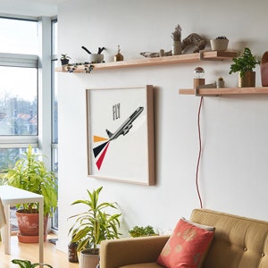 May include: A living room interior with a white wall, a large window, a white table with two chairs, a couch, and three wooden shelves with various plants, books, and a lamp. The middle shelf has a framed print of a plane with the word "FLY" above it.