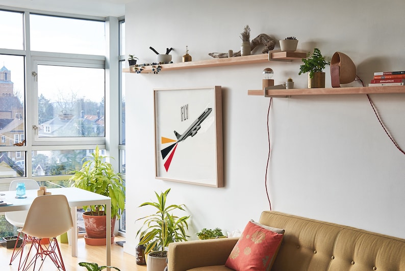 May include: A living room interior with a white wall, a large window, a white table with two chairs, a couch, and three wooden shelves with various plants, books, and a lamp. The middle shelf has a framed print of a plane with the word "FLY" above it.