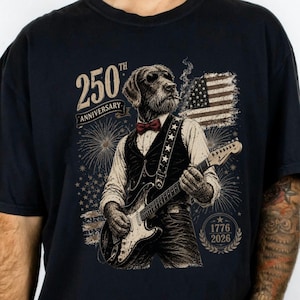 May include: A black t-shirt featuring a graphic of a dog wearing a vest and bowtie while playing an electric guitar. The design includes the text 250th Anniversary, 1776, 2026, a stylized American flag, and fireworks in shades of white, red, and gold.