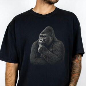 May include: A black short-sleeved t-shirt featuring a detailed graphic print of a gorilla in a contemplative pose, with its hand resting under its chin. The gorilla is rendered in shades of grey and black, centred on the chest of the shirt.