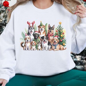 Peut inclure: Sweat-shirt blanc avec une illustration à l'aquarelle de plusieurs races de chiens ornées d'accessoires de vacances festifs. Les chiens sont regroupés autour d'un petit sapin de Noël. Le sweat-shirt est de style décontracté et confortable, idéal pour les fêtes.