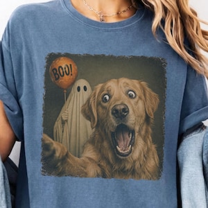 May include: A blue denim-colored t-shirt featuring a graphic print of a golden retriever with an open mouth and wide eyes, standing next to a ghost holding an orange balloon with the text BOO! printed in black on the balloon surface.