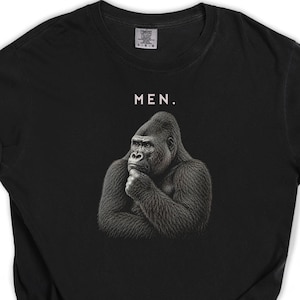 Puede incluir: Una camiseta negra de manga corta con un gráfico detallado de un gorila apoyando la barbilla en la mano en una pose pensativa. Sobre el gorila, la palabra "MEN." está impresa en letras mayúsculas blancas. Cuello redondo.