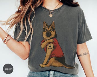 May include: A charcoal gray short-sleeve t-shirt featuring a graphic of a German Shepherd dog wearing a red t-shirt. The dog has a tattoo on its arm that reads I heart MOM with a heart symbol. The shirt has a crew neckline and a relaxed fit.