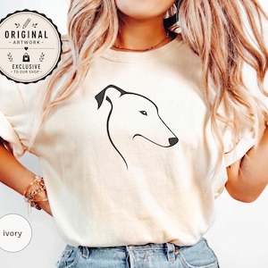May include: An ivory t-shirt featuring a minimalist black line art illustration of a greyhound dog profile. The shirt has a round neckline and short sleeves. A circular badge in the top left corner contains the text ORIGINAL ARTWORK EXCLUSIVE TO OUR SHOP.
