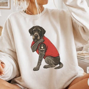 Könnte beinhalten: Ein sandfarbenes Sweatshirt mit Rundhalsausschnitt und einer grafischen Illustration eines dunkelhaarigen Deutsch Drahthaar-Hundes, der ein rotes T-Shirt trägt. Der Hund hat ein Tattoo am Oberarm mit der Aufschrift I heart MOM und einem Herz.