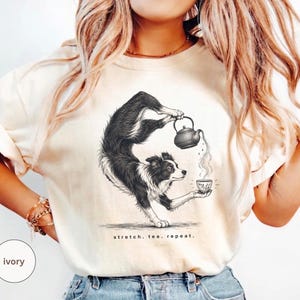 May include: An ivory t-shirt featuring a black and white graphic illustration of a border collie in a yoga stretch pose, holding a teapot and pouring tea into a cup. The text below the dog reads, stretch. tea. repeat. The shirt has short sleeves.
