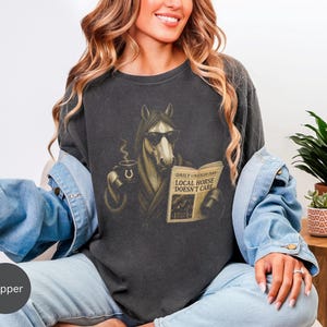 Vintage engraved horse wearing sunglasses and a robe while reading a newspaper and holding a steaming coffee mug. Humorous horse lover design featuring the headline “Local Horse Doesn't Care” in an antique newspaper style. Detailed equestrian artwork with a relaxed, unbothered attitude.