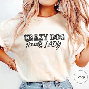 May include: Ivory-colored t-shirt with the text "CRAZY DOG LADY" in a distressed font. A black and white spotted bone graphic is between the words. The shirt is a crew neck style.