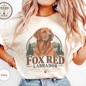 Peut inclure: Un t-shirt ivoire avec un motif central représentant un chien Labrador rouge renard dans un cadre circulaire orné de sapins et de montagnes. Le texte indique FOX RED LABRADOR et RARE SHADE, CLASSIC LAB HEART, avec une petite empreinte de patte.
