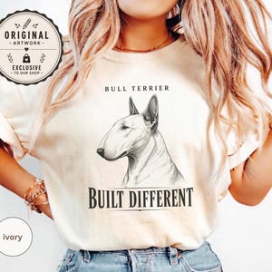 May include: An ivory t-shirt featuring a black and white sketch of a Bull Terrier dog head. Above the dog, the text reads BULL TERRIER, and below the dog, the text reads BUILT DIFFERENT. A circular badge in the top left corner says ORIGINAL ARTWORK.