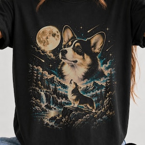 May include: A black t-shirt featuring a graphic print of a Corgi dog. The design includes a large Corgi head in the night sky, a smaller Corgi standing on a rocky cliff, a full moon, shooting stars, a waterfall, and a forest landscape.