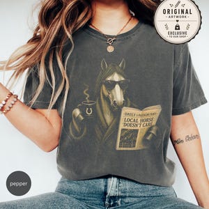 Vintage engraved horse wearing sunglasses and a robe while reading a newspaper and holding a steaming coffee mug. Humorous horse lover design featuring the headline “Local Horse Doesn't Care” in an antique newspaper style. Detailed equestrian artwork with a relaxed, unbothered attitude.