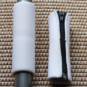 Könnte beinhalten: Zwei weiße Neopren-Griffbezüge. Einer ist um eine graue Metallstange gewickelt. Der andere hat einen schwarzen Reißverschluss in der Mitte. Die Bezüge dienen zum Schutz und zur Polsterung von Griffen.