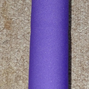 Puede incluir: Primer plano de un objeto cilíndrico con una funda de espuma morada. La funda cubre la parte superior del cilindro. El fondo es una superficie texturizada de color marrón claro.