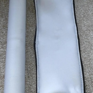 May include: A white padded sleeve with a black zip next to a white cylindrical object. The sleeve is rectangular and appears to be made of a soft, padded material. The zip runs along the edge of the sleeve. The cylindrical object is also white.