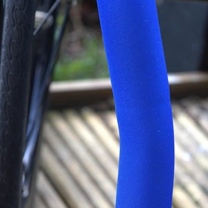May include: A close-up of a bicycle frame featuring a bright blue foam sleeve wrapped around a metal bar. The sleeve has a textured surface and curves gently. The background is blurred, with a bicycle wheel visible.