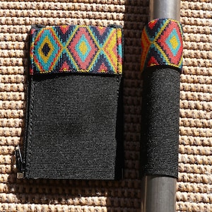 May include: A black elastic pouch with a zip and a colourful woven diamond pattern band at the top, alongside a matching black elastic band wrapped around a cylindrical object, both featuring red, yellow, and teal geometric designs.